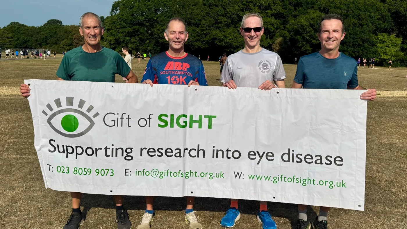 Four runners in running kit holding a white Gift of Sight banner reading Supporting research into eye disease, on a grass field with trees behind
