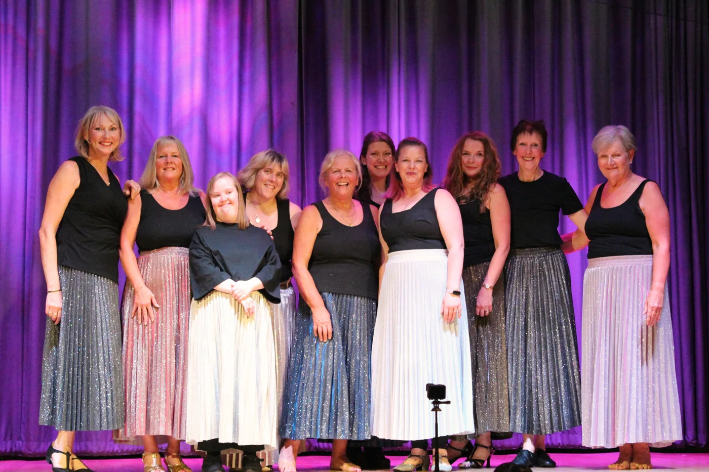 Ten performers in black tops and long pleated metallic skirts in silver, pink and white, standing together and smiling in front of a purple curtain